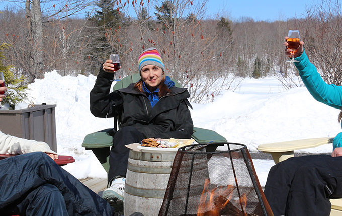 three women sitting in muskoka chairs raising wine glasses sitting by a fire on a sunny winter day