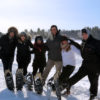 six people with arms linked raising one snowshoed foot against a winter background
