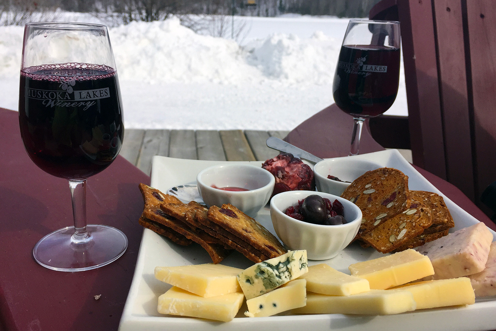 cheese plate with two glasses of muskoka lakes wine on the arm of Muskoka chairs against a snowy background