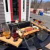 Two wine and cider tasting flights with a tabletop cheese roasting setup and a charcuterie board featuring crackers, meats, and cheeses, arranged on an outdoor glass patio table.