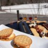 Close-up of two savory s’mores made with crackers, brie cheese, prosciutto, and preserves, with a blurred background showing a wine and cider tasting flight, charcuterie board, and snowy field under a blue sky.